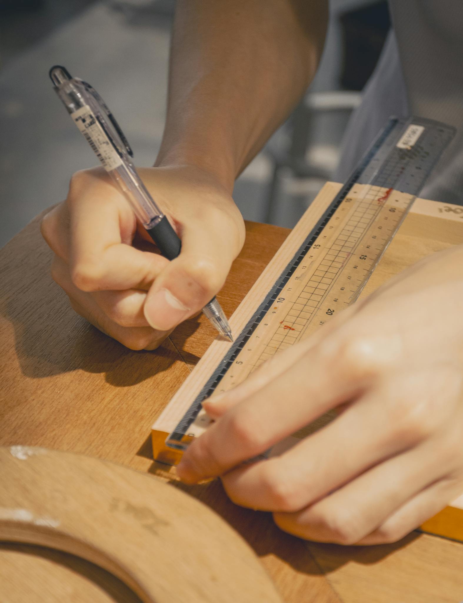 Close-up of hands using a ruler and pen on wooden surface in a well-lit workspace.