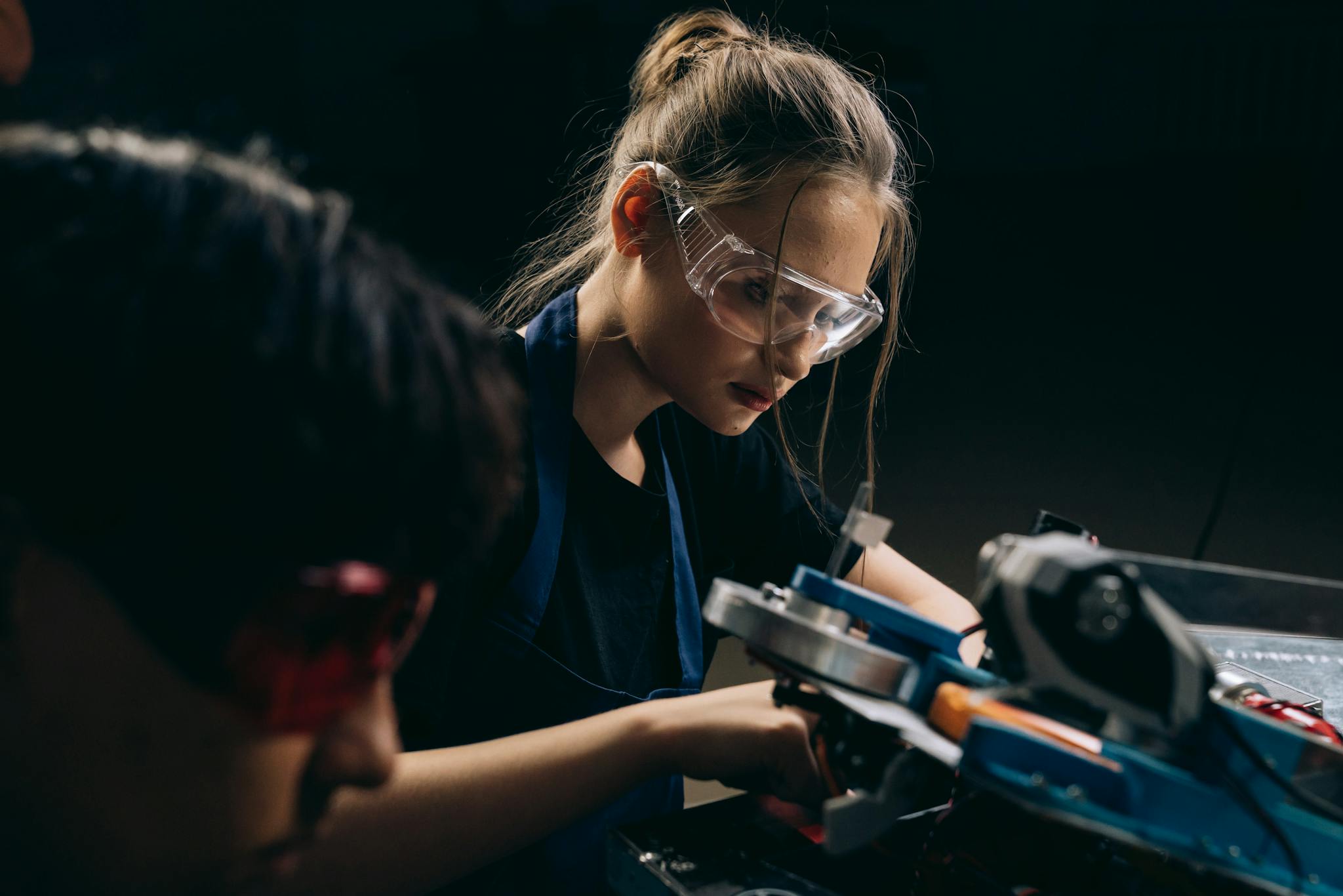 Two young adults in a workshop focused on a mechanical project, wearing protective goggles.